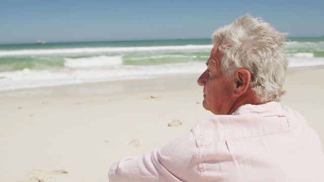 Senior Caucasian man sitting on the beach facing the ocean. Ideal for concepts related to peaceful living, retirement lifestyle, vacation, and serene nature settings. Can be used for promoting travel agencies, retirement planning services, or calming lifestyle articles.