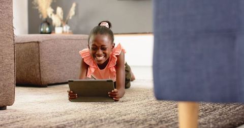 Joyful african girl lying on rug using tablet at home wearing coral ruffled top, playful learning