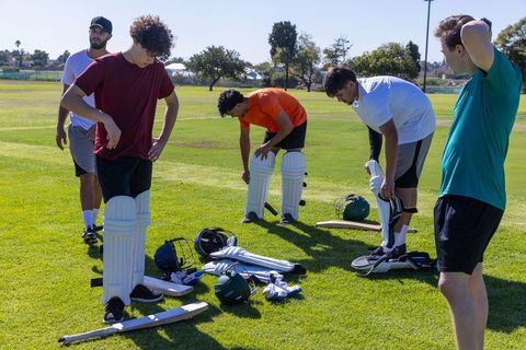 Multicultural men preparing for cricket practice on sunny park field with protective gear