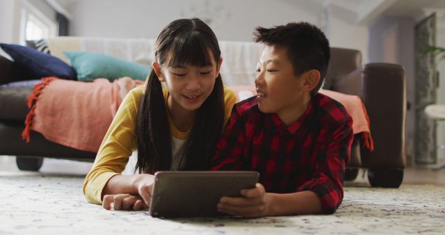 Two young Asian children lying on the floor in their living room, sharing and using a digital tablet. They are smiling and seem to be enjoying their time together. This image can be used for promoting family activities, digital learning, sibling bonding, and responsible screen time management.