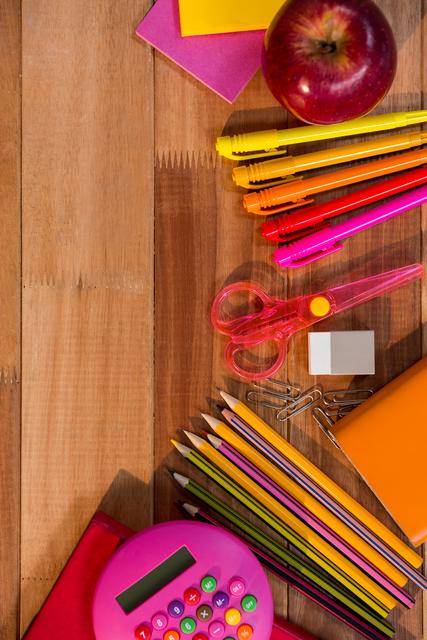 Colorful school supplies neatly arranged on a wooden table. Items include pencils, pens, a calculator, scissors, an apple, an eraser, and paper clips. Ideal for back-to-school promotions, educational materials, classroom decor, and office supply advertisements.
