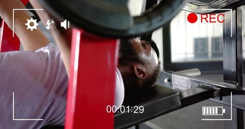 Man performing heavy bench press recorded with on-screen camera overlay in gym