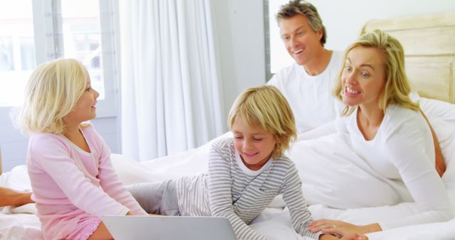 A Caucasian family enjoys time together in a bright bedroom, with copy space. Smiling parents watch their children, a boy and a girl, use a laptop while sitting on the bed, creating a warm, domestic scene.