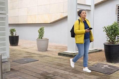 Woman walking in yellow jacket with backpack and smartphone in modern urban courtyard