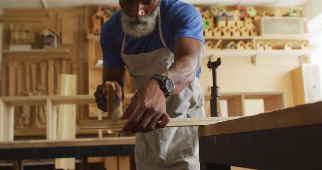 Senior craftsman sanding wooden plank in a workshop environment, using carpentry tools. He wears an apron and works with precision and focus on his task. Ideal for illustrating concepts of craftsmanship, woodworking skills, traditional trades, aging workforce, and pursuing hobbies in senior years.