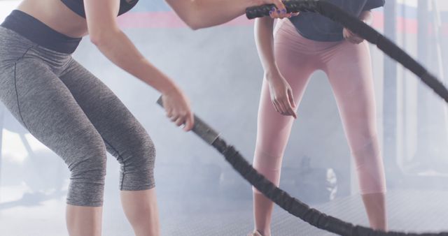 Image of diverse female fitness trainer with stopwatch and woman battling ropes working out at a gym. Exercise, fitness and healthy lifestyle.