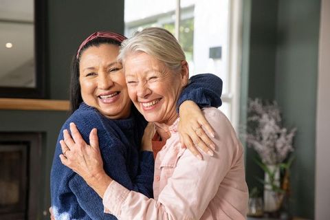 Mature women embracing and laughing by window in cozy home friendship moment