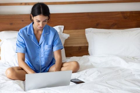 Woman wearing blue satin pajamas working on laptop while sitting cross-legged on bed