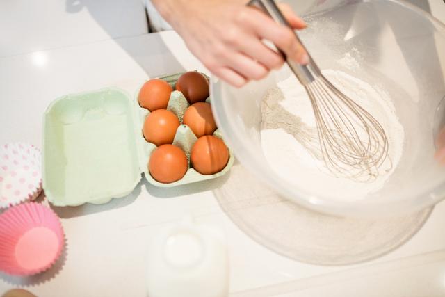 This image shows a woman whisking flour in a bowl, with eggs, milk, and cupcake liners nearby. Ideal for use in cooking blogs, recipe websites, culinary magazines, and advertisements for kitchenware or baking supplies.