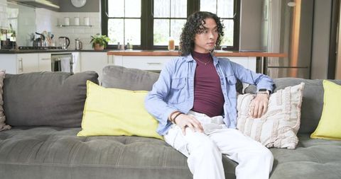 Non-binary person relaxing on modern gray sofa in bright open-plan kitchen with yellow pillows
