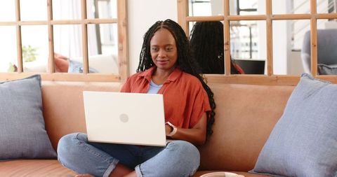 African woman working on laptop on cozy sofa at home, remote work lifestyle