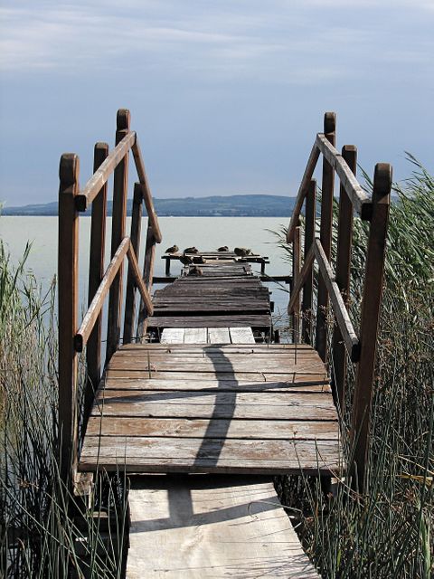 Old wooden dock extends into calm lake, framed by rustic railings. Sky meets water in tranquil scene, making it perfect for nature or travel blogs, serene lake-oriented projects, relaxation theme prints, or outdoor recreational advertisements.