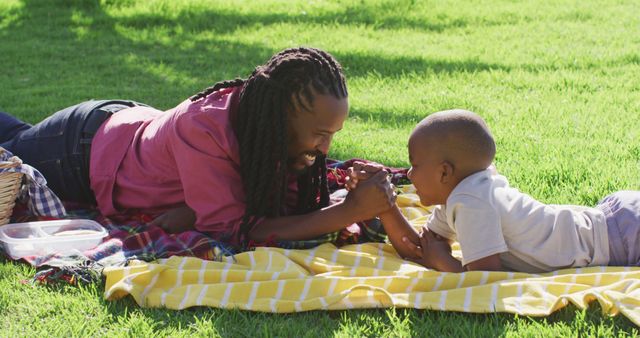 Image of happy african american father and son having picnic on grass, arm wrestling. family, togetherness, spending quality time together outdoors.