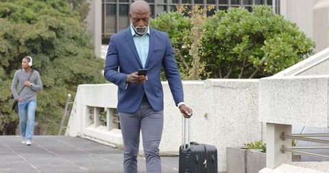 Mature african american businessman walking with rolling suitcase while checking smartphone