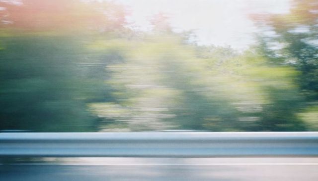 Motion-blurred highway guardrail with sunlit treeline streaks, pastel color wash, roadside motion