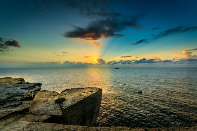 Vibrant sunset over calm ocean viewed from high, rocky cliff. Sun rays break through scattered clouds, casting beautiful glow on water. Perfect for travel brochures, nature photography projects, and relaxation-themed content.