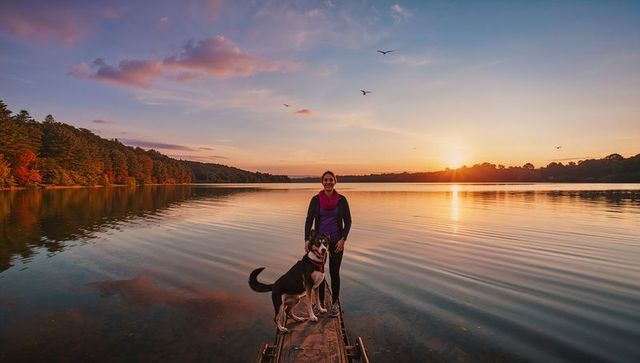 Woman and dog watching autumn sunset on lake dock at golden hour