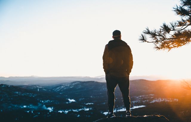 A man stands on a mountain peak, observing a breathtaking sunrise over a vast scenic landscape. Ideal for themes like adventure travel, exploration, achieving goals, enjoying nature, outdoor activities, and finding peace.