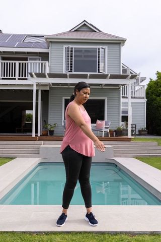 Mature woman standing at plunge pool edge in modern backyard wearing pink top and black leggings