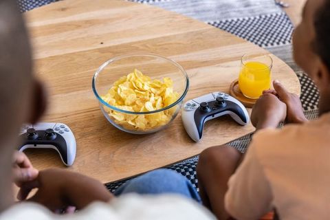 African american father and son playing games with wireless controllers, chips and juice