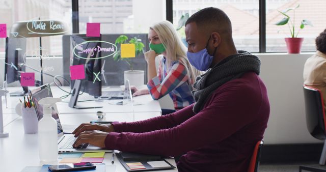 Professionals sitting at desks, using laptops while wearing masks during the pandemic, displaying adherence to safety protocols. This stock photo is ideal for illustrating modern office environments, business culture, remote work, pandemic adaptations, and health and safety measures in workplaces.