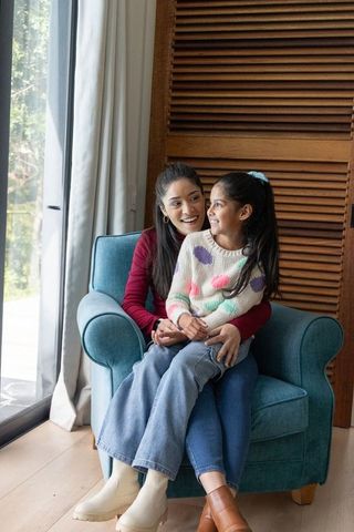 Indian mother and daughter cuddling on teal armchair by sunlit sliding door
