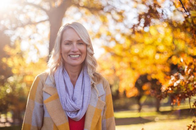 Woman standing in park during autumn, surrounded by colorful fall foliage. She is smiling and wearing a scarf and casual clothing. Ideal for use in lifestyle, seasonal, and outdoor leisure content.