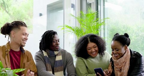 Diverse friends laughing and sharing smartphone content on cozy livingroom couch with scarves