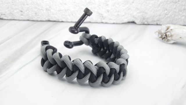 Braided paracord bracelet featuring black and gray cords placed on white marble surface with open clasp displayed. Suitable for use in jewelry collections, outdoor gear promotions, or fashion accessory catalogs. Perfect for marketing adventure, survival gear, or handmade craft products.