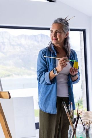 Senior woman painting in sunlit lakeside studio with easel, brushes and yellow paint
