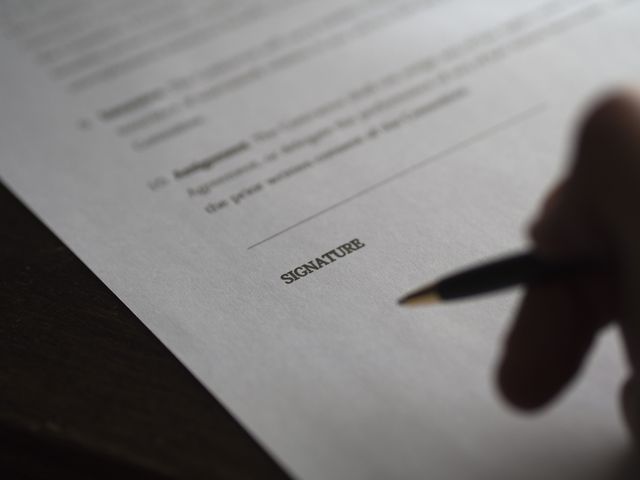 Close-up shows a hand holding a pen about to sign contract documentation. Useful image for illustrating legal agreements, business deals, member enrollments, or financial transactions.