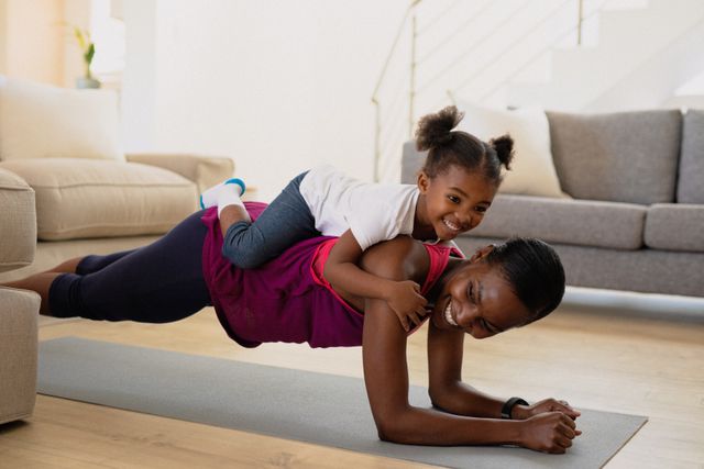 This image shows a joyful African American mother and daughter engaging in a playful exercise session at home. The mother is performing a plank while the daughter is lying on her back, both smiling and enjoying their time together. This image is perfect for promoting family fitness, healthy lifestyles, and the importance of spending quality time with loved ones. It can be used in articles, advertisements, and social media posts related to family activities, home workouts, and wellness.