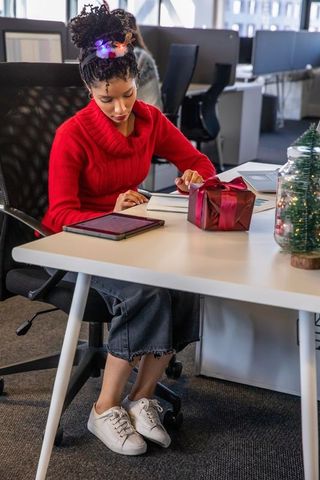 African american woman writing in planner at office desk with wrapped gift and lights