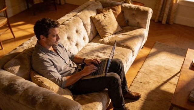 Man working on laptop on tufted sofa in sunlit cozy living room, remote work productivity