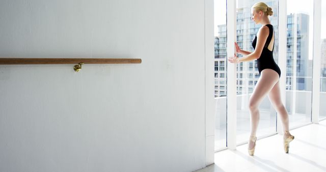 Ballerina practicing ballet positions in an urban dance studio, wearing black leotard and pointe shoes, demonstrating poise and balance. This image is ideal for use in dance studio promotions, ballet schools, articles on performing arts, and advertisements related to ballet clothing or equipment.