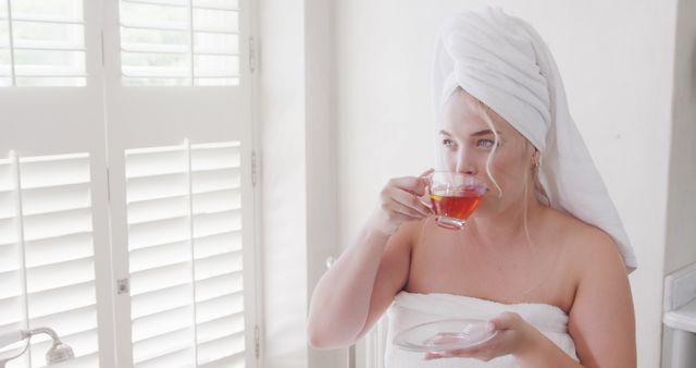 A woman is drinking tea in a bathroom, wrapped in a towel with her hair wrapped in another towel. Useful for themes related to relaxation, self-care, morning routines, and wellness at home visuals such as blog posts, social media content, or advertisements for spa services.