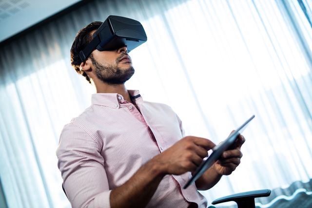 Businessman in an office setting, wearing a VR headset and interacting with a tablet. Ideal for use in articles, blogs, and presentations about modern technology, innovation in business, virtual reality applications, and the integration of digital tools in the workplace.