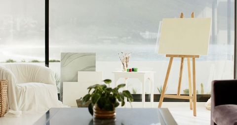 Sunlit waterfront art studio featuring blank canvas on wooden easel, paints and brushes