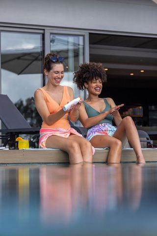 Two friends applying sunscreen at modern poolside with cocktail, smiling and relaxing