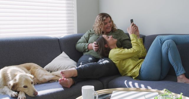 Caucasian lesbian couple smiling and sitting on couch with dog. domestic life, spending free time relaxing at home.