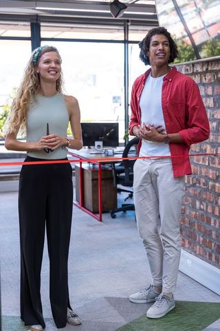 Diverse coworkers ceremoniously cutting red ribbon in modern office