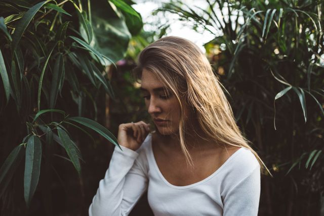 Young woman is standing amidst lush tropical foliage, appearing thoughtful and reflective. She is dressed in a casual white shirt. This content can evoke themes of tranquility and connection with nature, suitable for advertisements, wellness content, or lifestyle blogs.