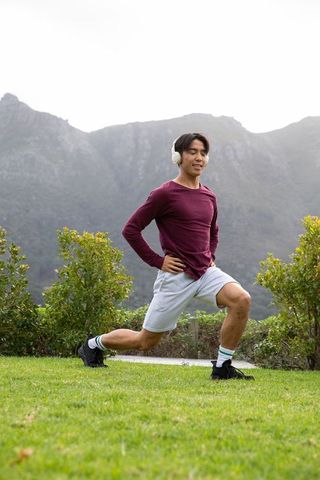 Young asian man doing forward lunge in headphones on mountain lawn for outdoor fitness