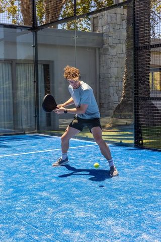 Young man playing padel on vivid blue court with glass walls and summer sunlight