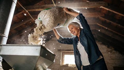 Senior woman pouring raw wool into hopper in rustic loft, sunlight backlighting dust and fibers