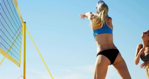 Female athlete spiking volleyball on sunny beach court with yellow net, teammate reacting