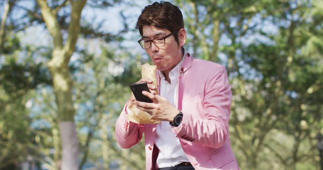 Man enjoying food while engaged with smartphone in sunny park. Helps in showcasing concepts of multitasking, modern lifestyle, and outdoor activities. Ideal for use in advertisements for food delivery services, tech products, or lifestyle blogs.