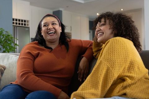 Diverse female friends laughing on couch wearing warm sweaters, enjoying cozy chat in living room