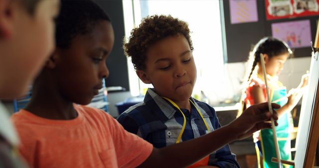 Diverse group of children painting on easels in a classroom, exploring their creativity in an art lesson. Ideal for topics related to education, childhood development, creative learning environments, and multicultural classrooms.