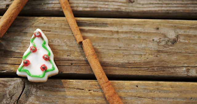 A Christmas tree-shaped cookie adorned with green icing and red decorations lies next to cinnamon sticks on a rustic wooden surface, with copy space. The festive arrangement evokes the warmth of holiday baking and traditions.